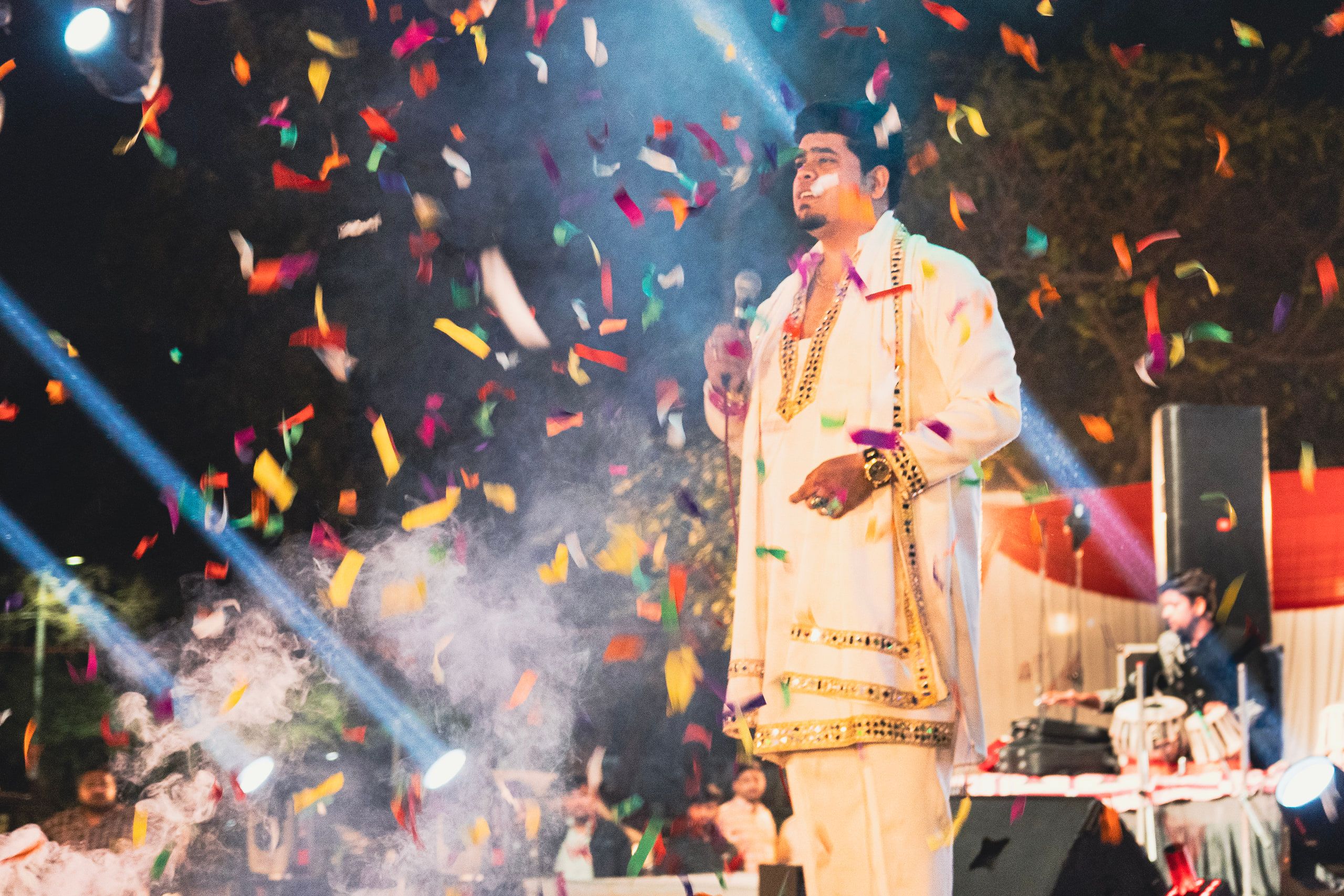 A performer in a white sherwani on stage as confetti and smoke fill the air