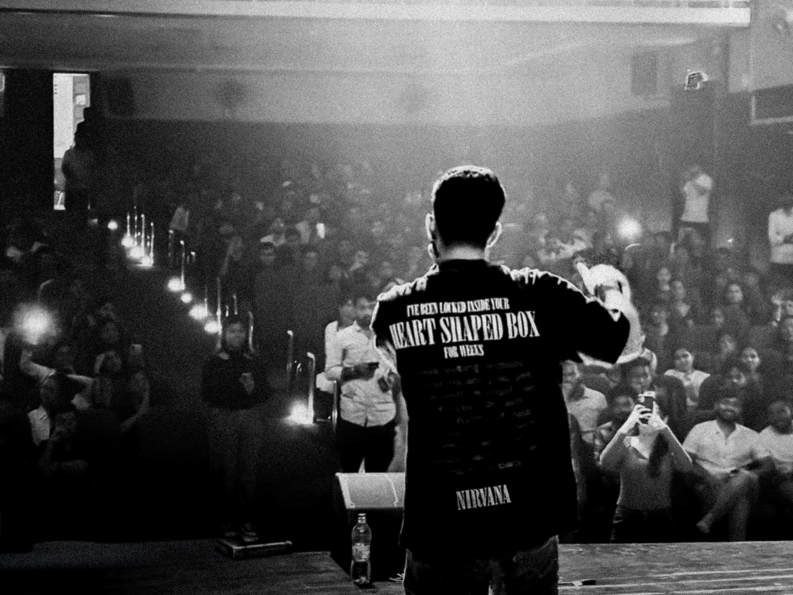 Black and white shot from behind a performer looking out at a packed hall lit by phone torches